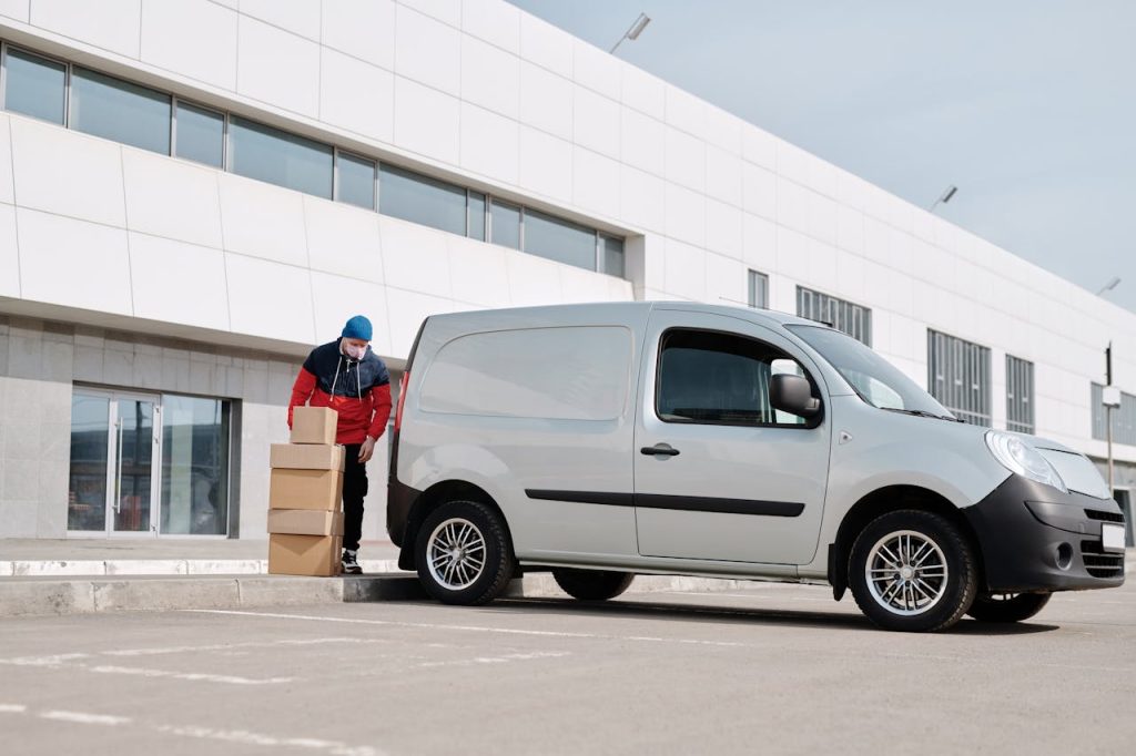 delivery-man-with-boxes-next-to-a-white-van-4391470 A delivery driver wearing a face mask unloads packages from a van outside a modern building.